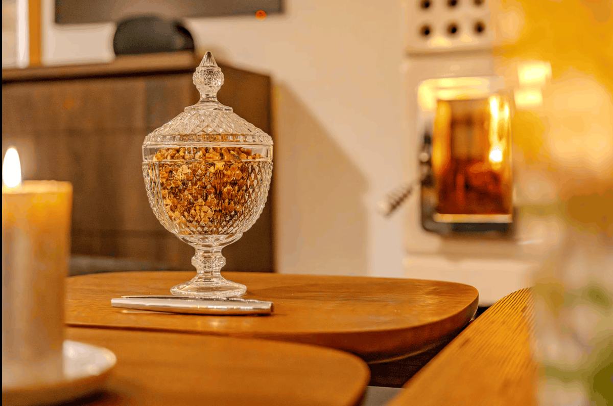 A clear crystal glass jar with a lid, filled with dried botanicals, displayed on a wooden table in a cozy indoor setting with a candle and warm light in the background.