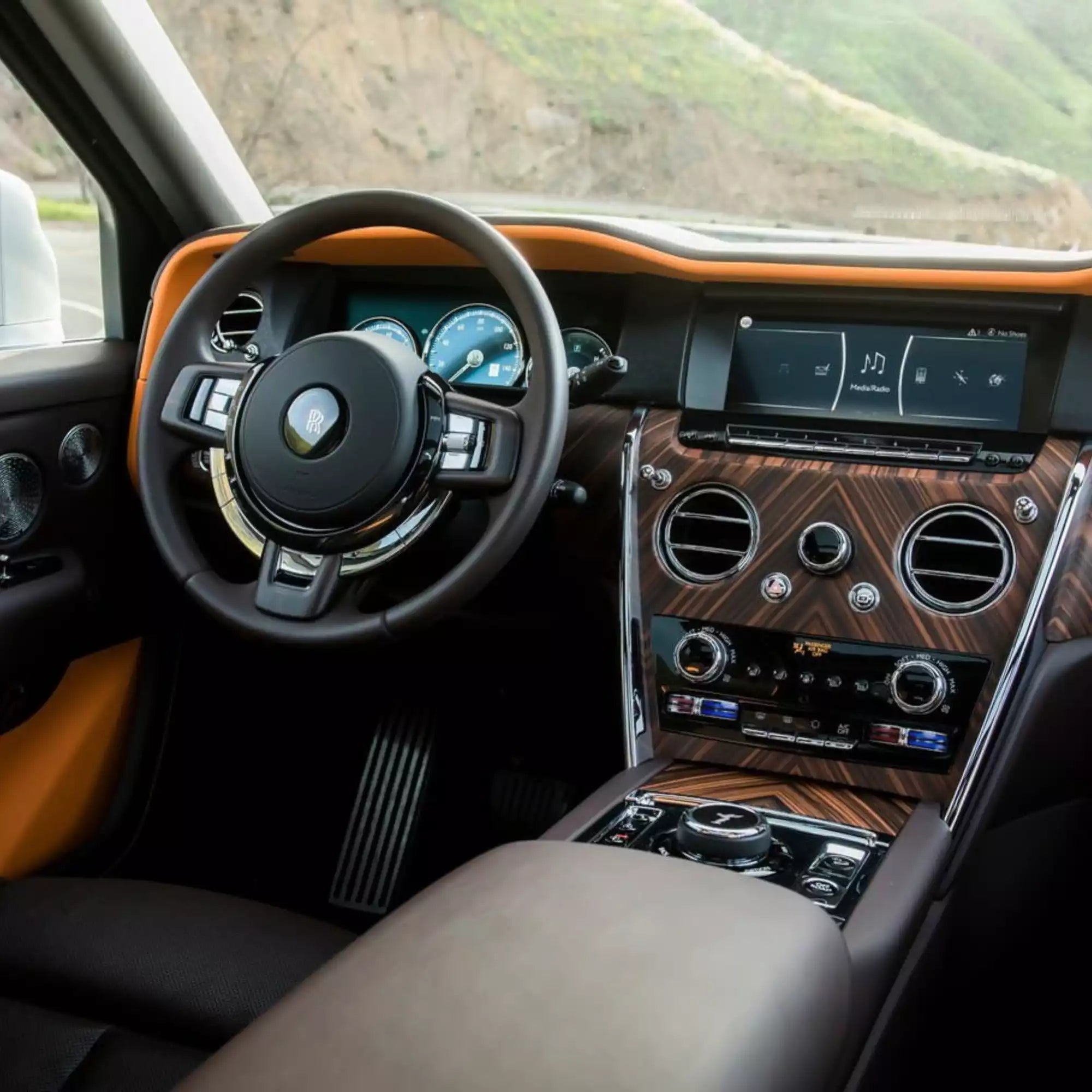 Luxury car interior featuring a black leather steering wheel with a gold crest, wood trim on the dashboard and center console, and bright orange accent panels, shown with a blurred outdoor background.