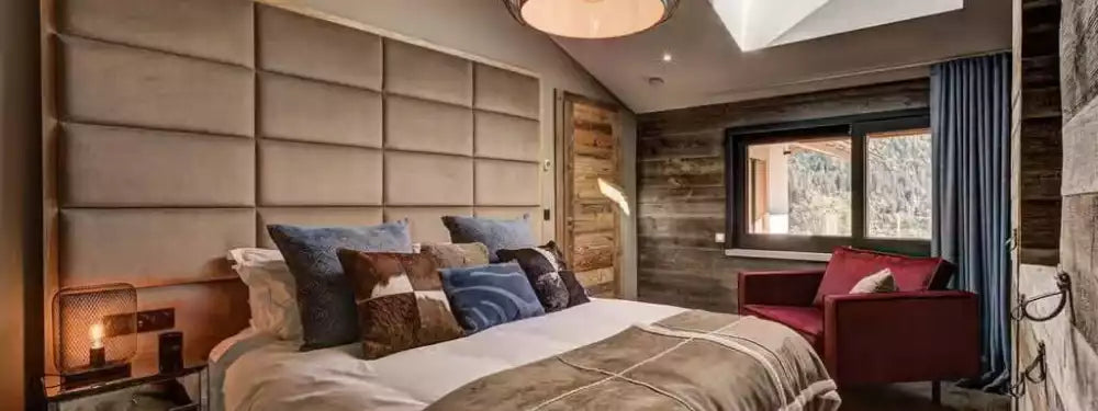 A modern bedroom interior featuring a large bed with beige and blue pillows, a tall padded headboard, a red armchair by a window, and wood-paneled walls.