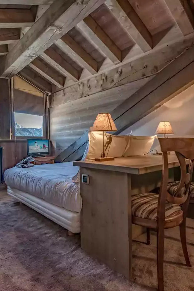 A rustic attic bedroom with exposed wooden beams, a bed with white bedding, a small wooden desk with two chairs, and warm lamps, photographed indoors.