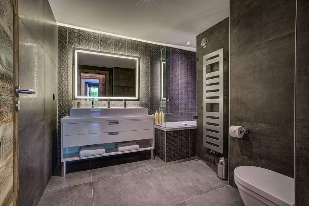 Modern bathroom vanity area with a white double-sink console, large backlit mirror, dark patterned wall tiles, gray floor tiles, and a white heated towel rail.
