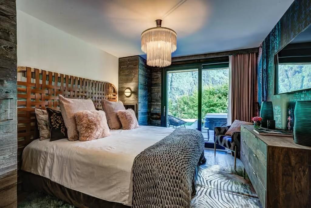 A styled bedroom interior featuring a large bed with cream bedding, textured pink and brown pillows, a wooden lattice headboard, a hanging cylindrical chandelier, a patterned rug, and a dresser with green ceramic vases near tall windows overlooking a forest.