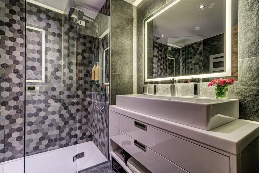 Modern bathroom vanity with a white vessel sink, chrome faucets, a large backlit mirror, and a glass shower enclosure featuring gray hexagonal mosaic tile walls.
