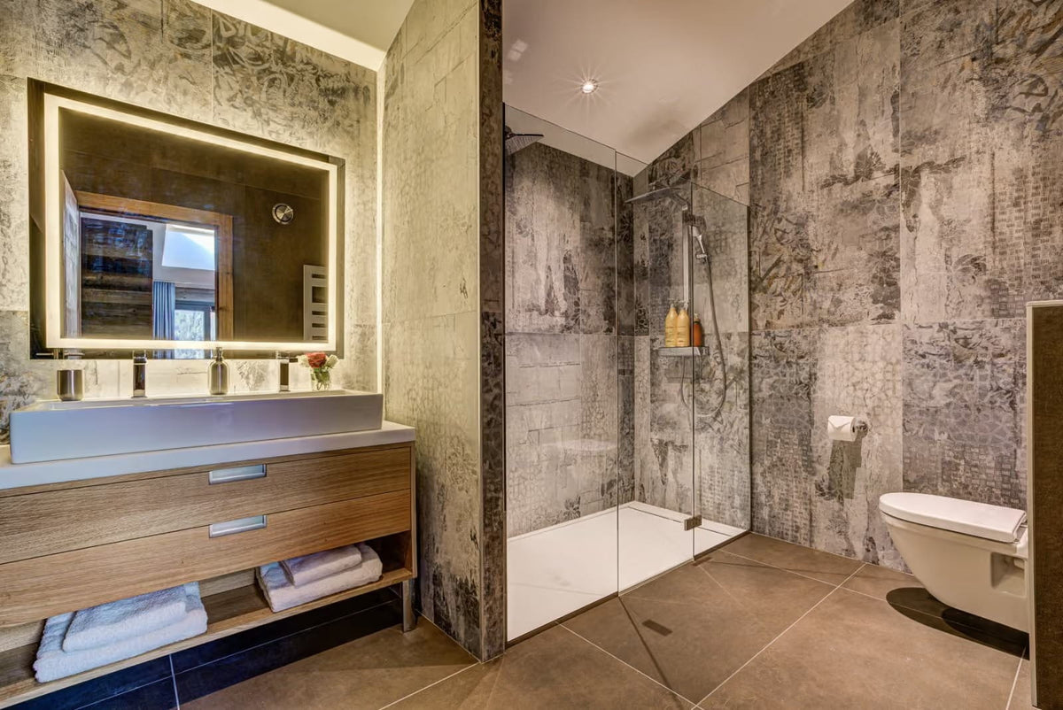 A modern bathroom interior with a large illuminated rectangular mirror, wood-and-slate vanity, walk-in shower with glass enclosure, tiled walls, and a white toilet. No people or animals are present.