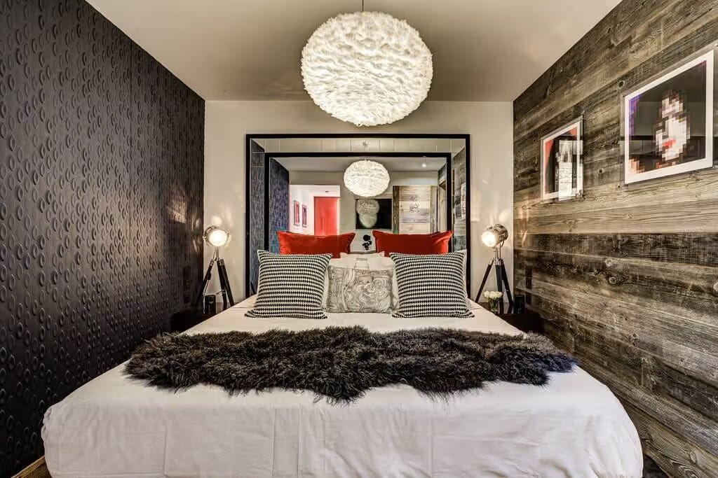 A modern bedroom featuring a large bed with white bedding, striped pillows, and a dark shag rug. The room has textured black wallpaper on one side, a rustic dark wood-paneled wall on the other, and a large black-framed mirror on the back wall reflecting a white textured chandelier.