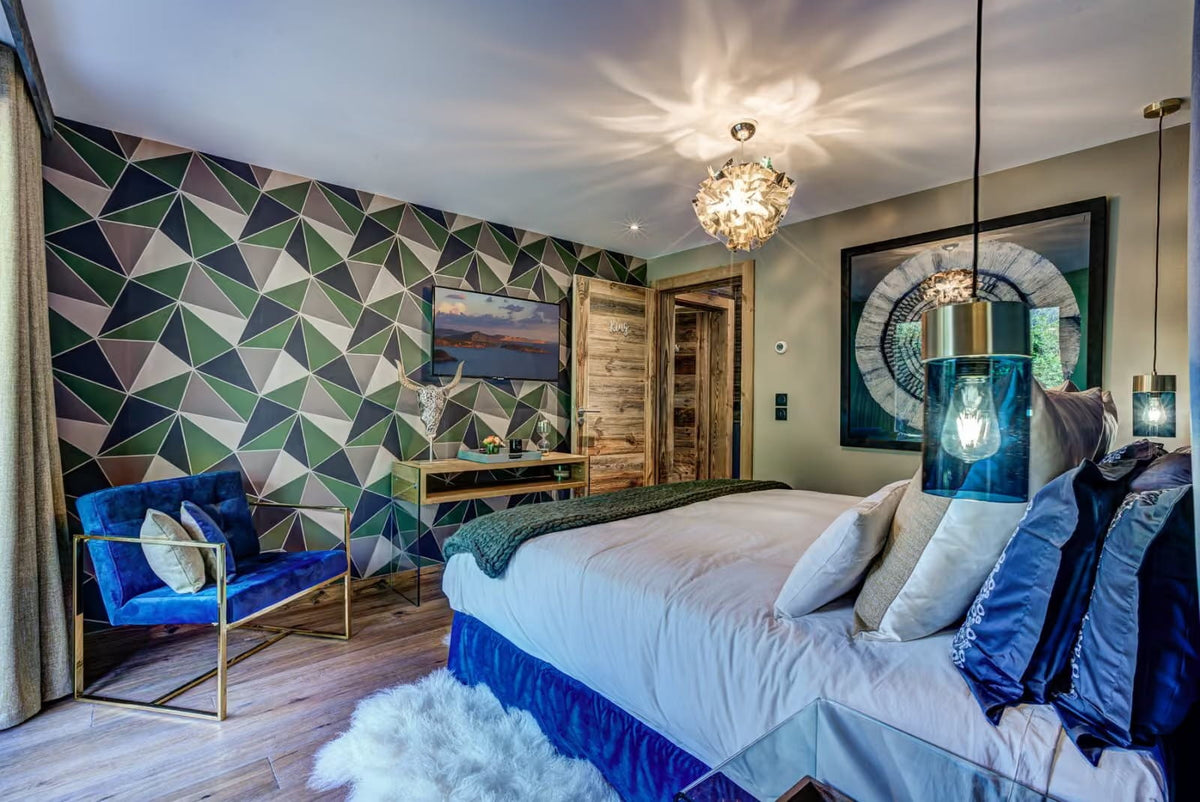 A modern bedroom interior featuring a bed with white bedding and blue pillows, a blue upholstered armchair with gold legs, geometric patterned wallpaper, wood flooring, and decorative pendant lights.