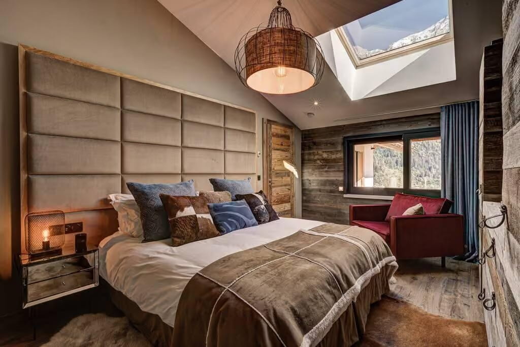 A luxury mountain-style bedroom featuring a large tufted beige headboard, a bed with brown and blue pillows, a red armchair, a rustic wood wall, and a skylight looking out to snowy peaks.