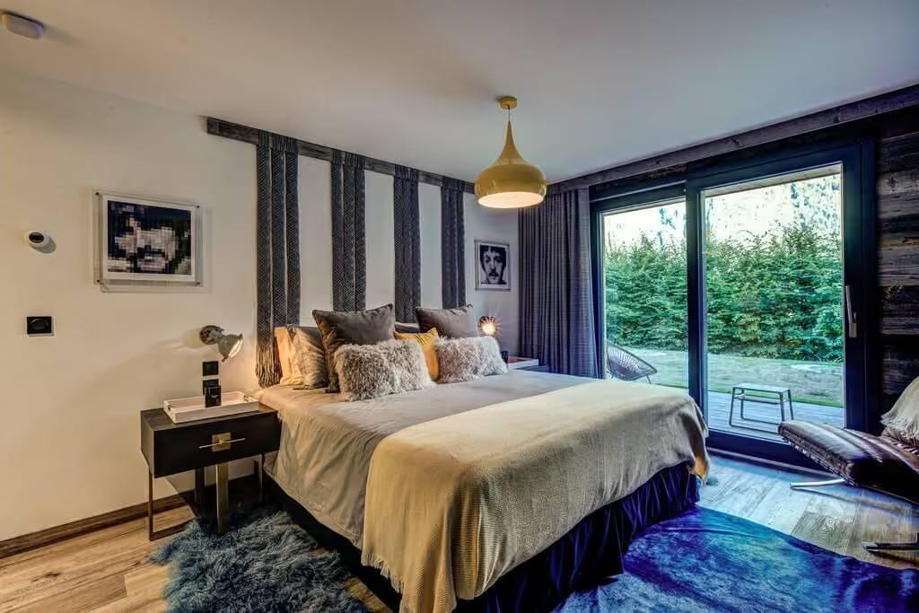A modern bedroom featuring a bed dressed in beige and gray bedding, styled with striped headboard panels, a blue shag rug, and large floor-to-ceiling windows overlooking a green garden.