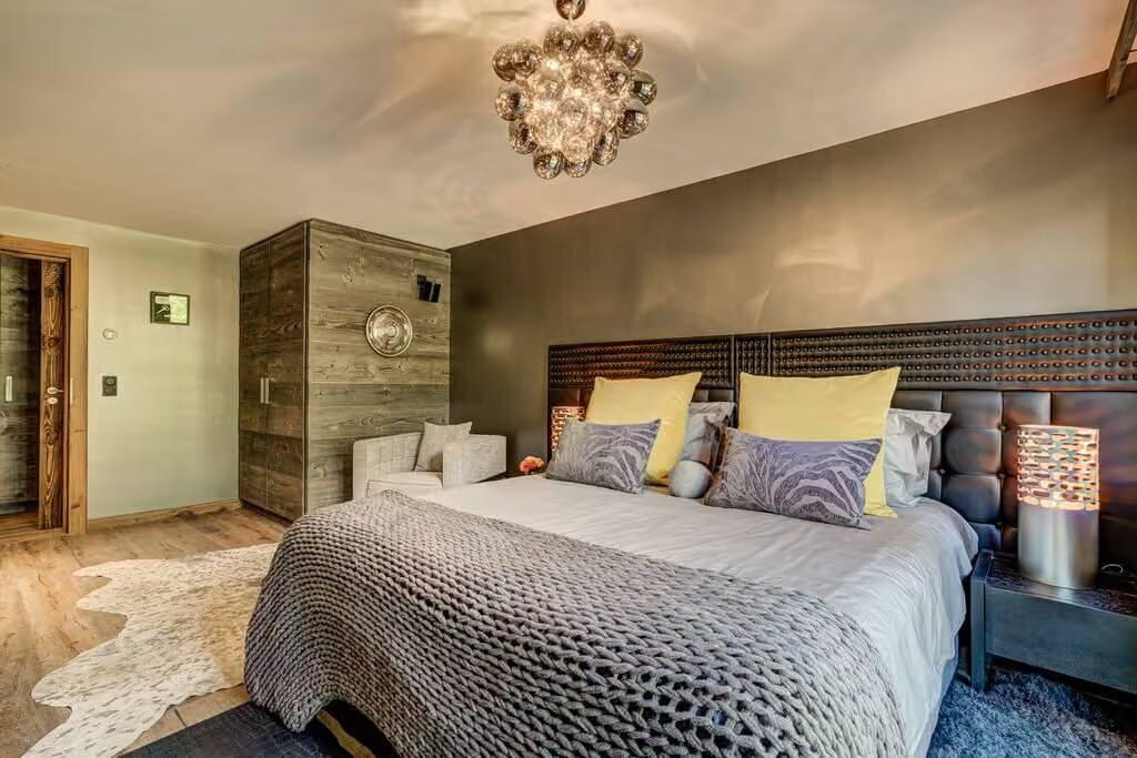 A modern bedroom featuring a large bed with a textured gray knit duvet, yellow and gray pillows, a dark upholstered headboard, and a decorative wood-paneled wall section behind it.