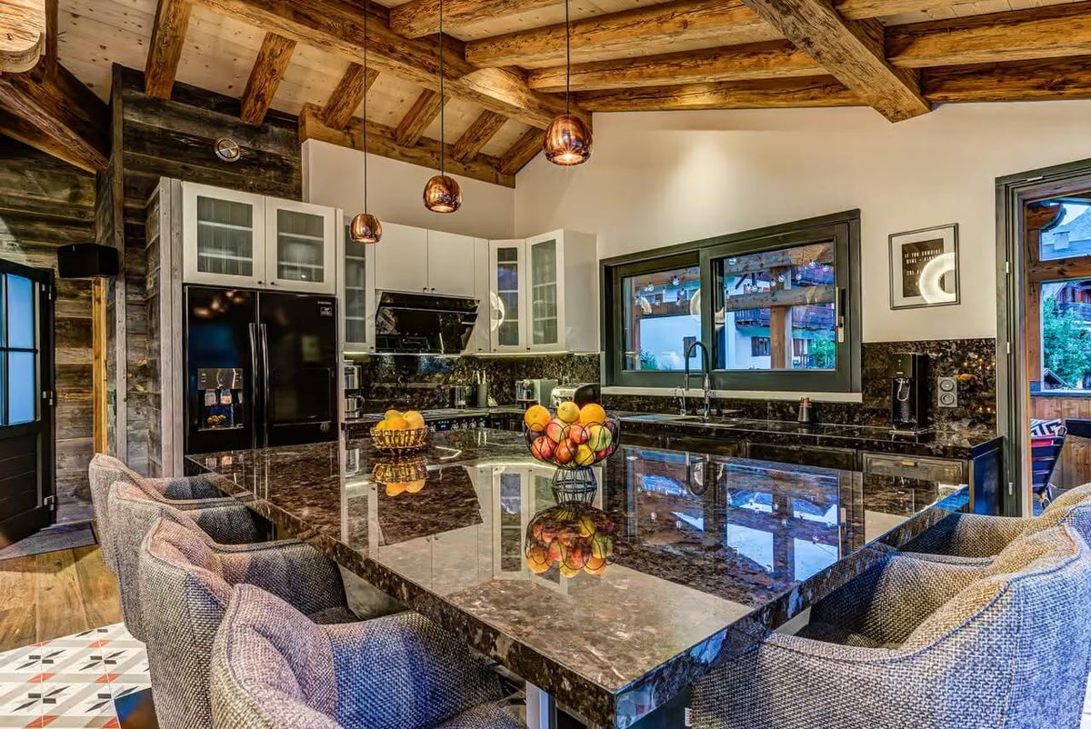 A luxury modern kitchen with a large dark marble island, textured gray upholstered bar stools, rustic wooden ceiling beams, and black stainless steel appliances.