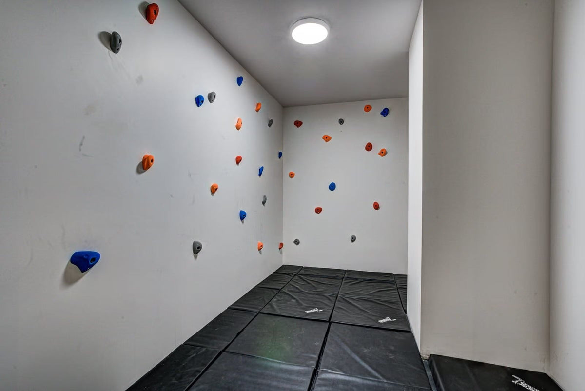 An indoor bouldering gym corner with white walls, multicolored climbing holds, and black padded floor mats under a ceiling light.