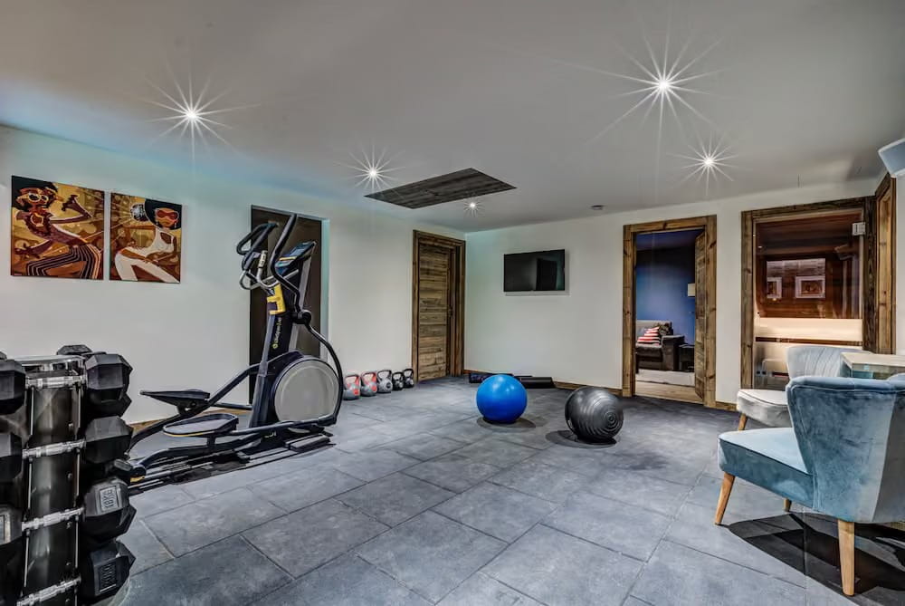 A spacious home gym interior with a gray stone floor, black elliptical machine, dumbbell rack, blue exercise ball, black ball, framed wall art, open wooden doors, and a blue upholstered chair in the foreground.
