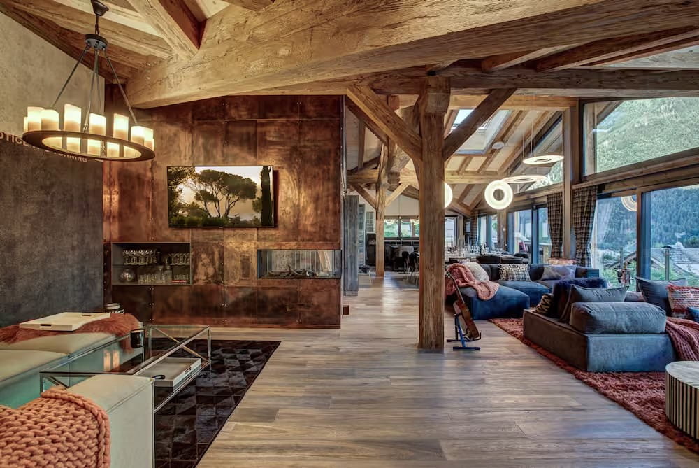 A luxurious mountain chalet living room featuring rustic wooden beams, a large copper-brown fireplace wall with a mounted landscape print, a modern chandelier, and plush blue sofas on patterned rugs.