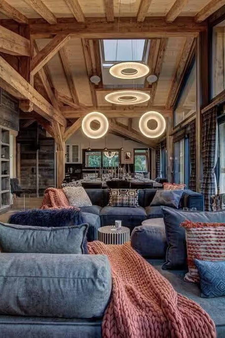 A rustic mountain chalet living room with exposed wooden beams, a large blue lounge chair in the foreground, patterned cushions, a pink throw blanket, and circular ceiling lights. Large windows offer a view of the outdoor mountain landscape.