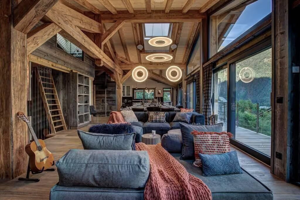 A rustic mountain chalet living room with exposed wooden beams, a large blue lounge chair in the foreground, patterned cushions, a pink throw blanket, and circular ceiling lights. Large windows offer a view of the outdoor mountain landscape.