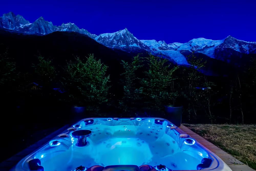A glowing blue outdoor hot tub set against a dark mountain landscape with snow-capped peaks under a deep blue night sky.