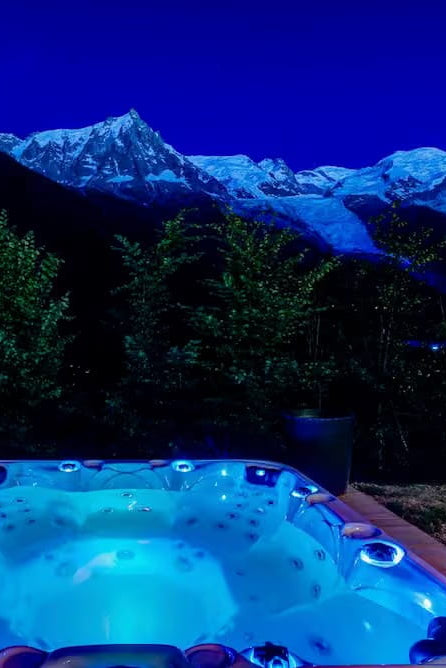 A glowing blue outdoor hot tub set against a dark mountain landscape with snow-capped peaks under a deep blue night sky.