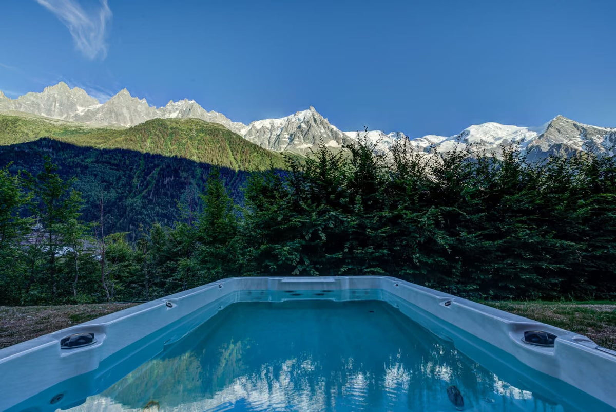 A rectangular outdoor hot tub with clear blue water in the foreground, set against a backdrop of dark green pine trees and majestic snow-capped mountains under a bright blue sky.