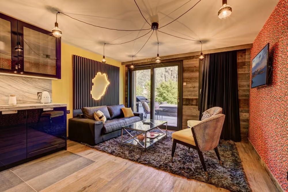 A modern living room interior with a dark gray sofa, glass coffee table, textured rug, wood flooring, yellow accent wall, patterned orange wall, and large windows with outdoor greenery visible.
