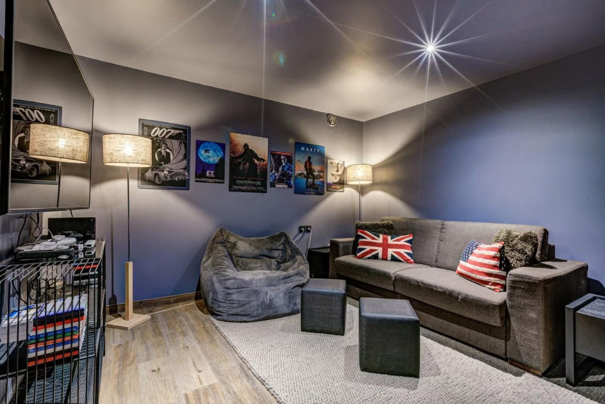 A modern home theater or lounge room with a gray fabric sofa decorated with Union Jack and American flag pillows, a matching gray beanbag chair, two dark gray ottomans, and movie posters on the wall.