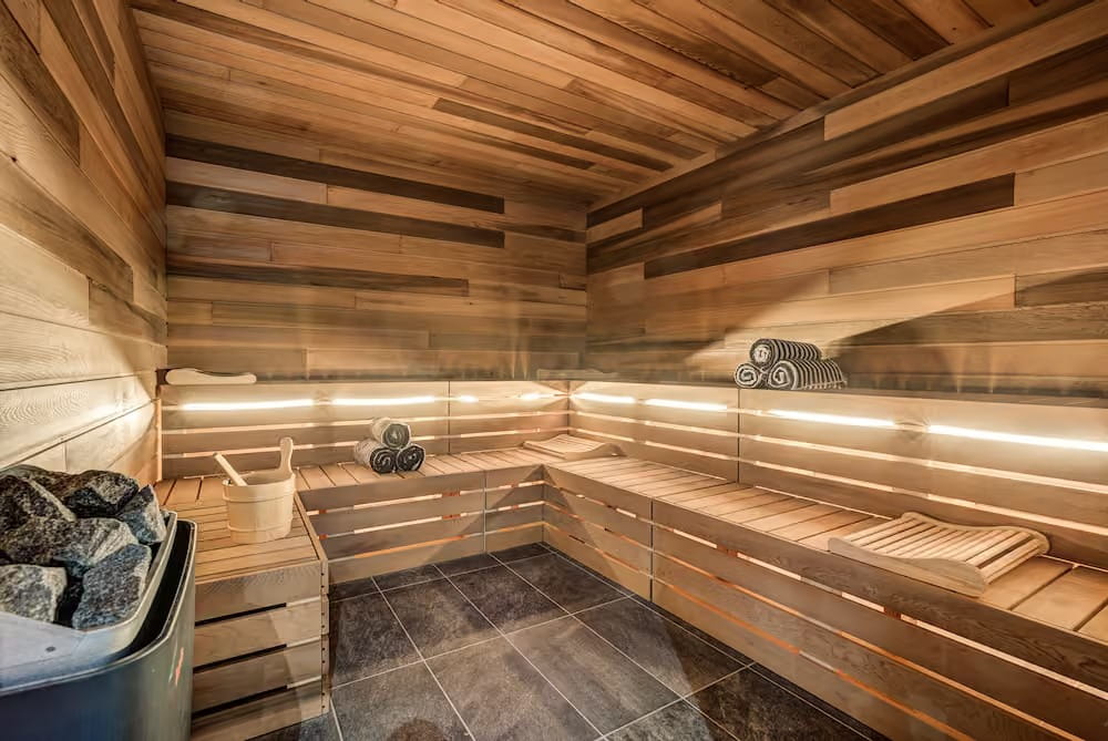 A wooden sauna room with tiered wooden benches, warm ambient lighting, a bucket of rocks, and a white bucket and ladle on the floor.
