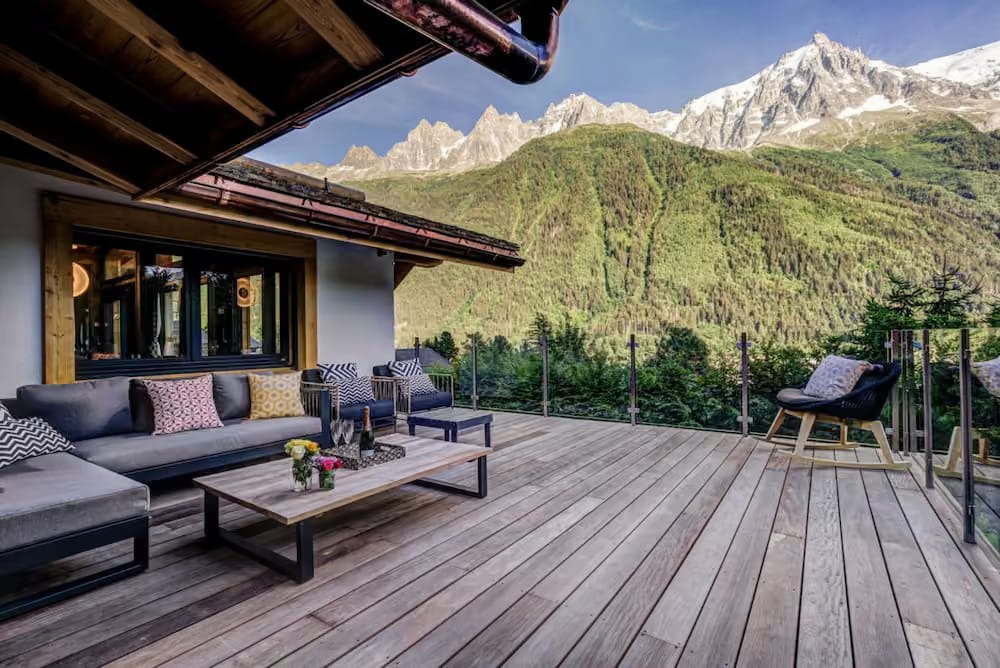 Modern outdoor patio furniture set on a wooden deck overlooking a lush green mountain valley and snow-capped peaks under a clear blue sky.