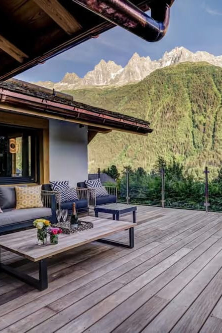 Modern outdoor patio furniture set on a wooden deck overlooking a lush green mountain valley and snow-capped peaks under a blue sky.
