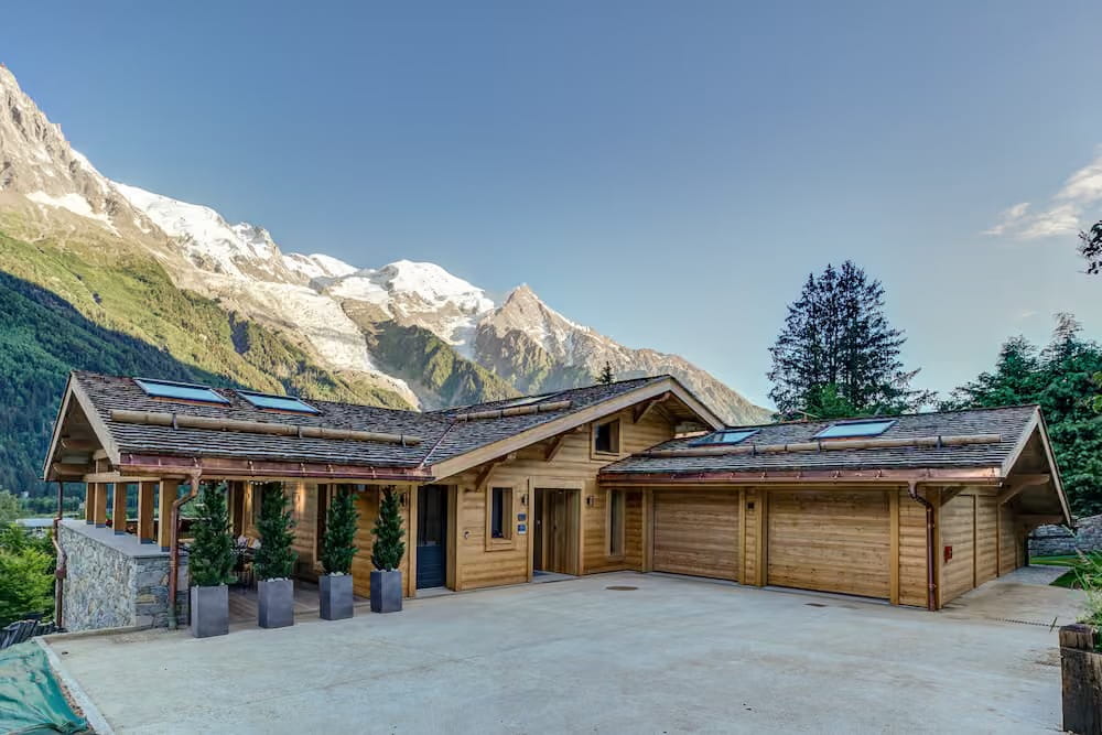 A modern wooden chalet-style house with a large concrete driveway, set against a backdrop of snow-capped mountains and a clear blue sky.