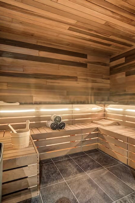 A wooden sauna room with tiered wooden benches, warm ambient lighting, a bucket of rocks, and a white bucket and ladle on the floor.