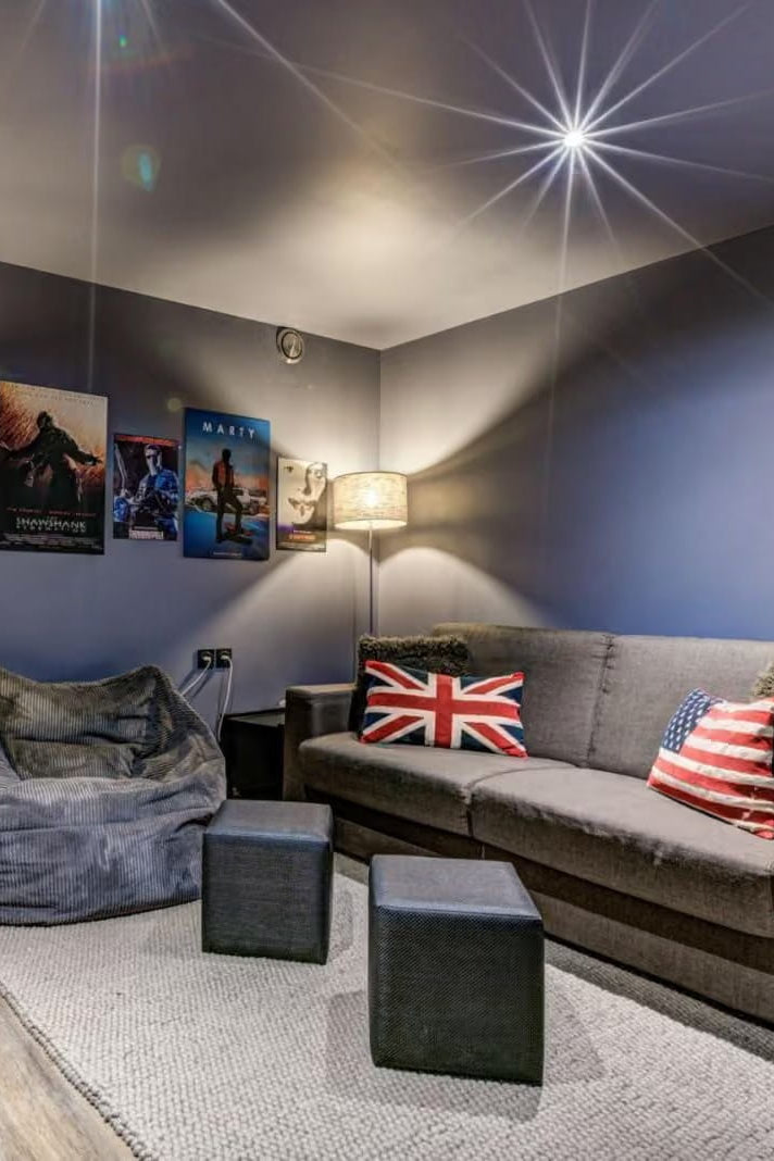 A modern home theater or lounge room with a gray fabric sofa decorated with Union Jack and American flag pillows, a matching gray beanbag chair, two dark gray ottomans, and movie posters on the wall.