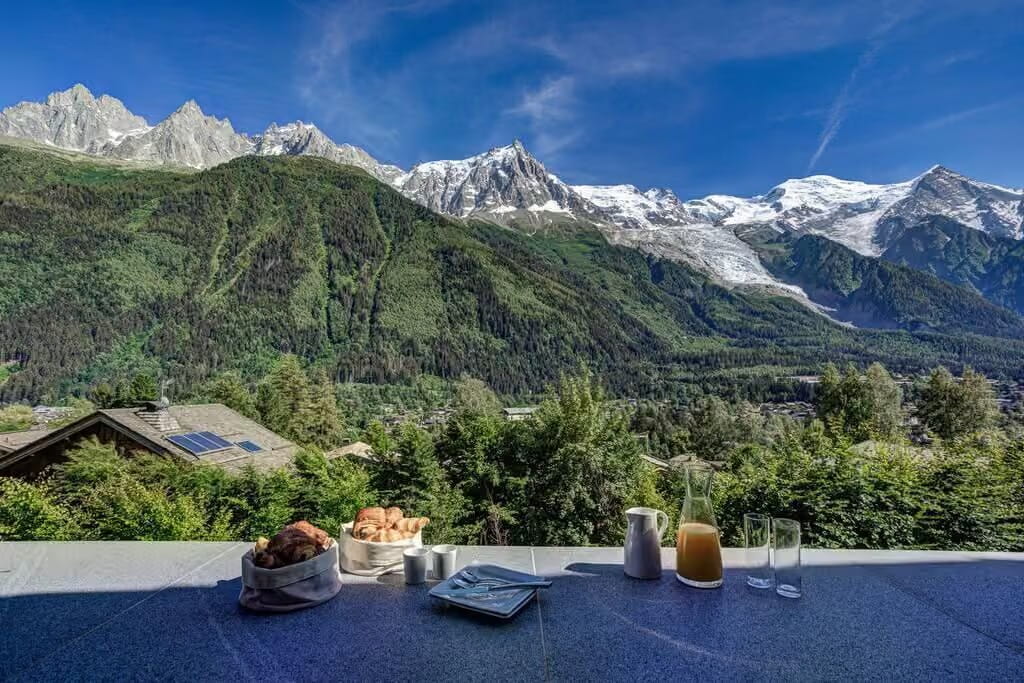 A scenic mountain landscape with snow-capped peaks, green slopes, and a blue sky, viewed from a balcony or terrace with a table setting in the foreground including bread, a pitcher of orange juice, and glasses.