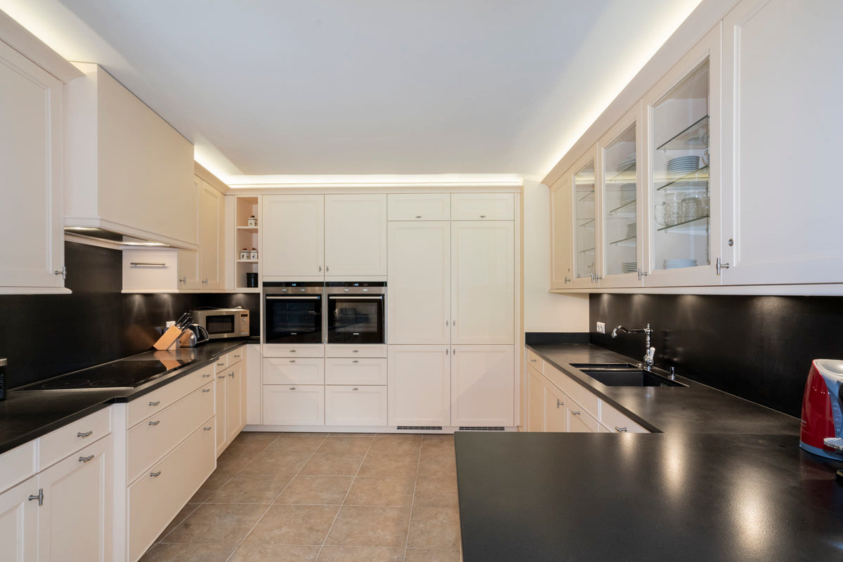 A modern kitchen interior with white cabinetry, black countertops and backsplash, tiled floor, built-in ovens, and glass-front upper cabinets.