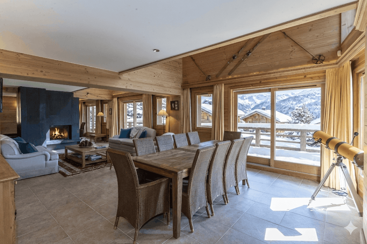 A bright, modern ski chalet dining and living room featuring a wooden dining table and chairs, a fireplace, large windows with a view of snowy mountains, and a telescope on a tripod.