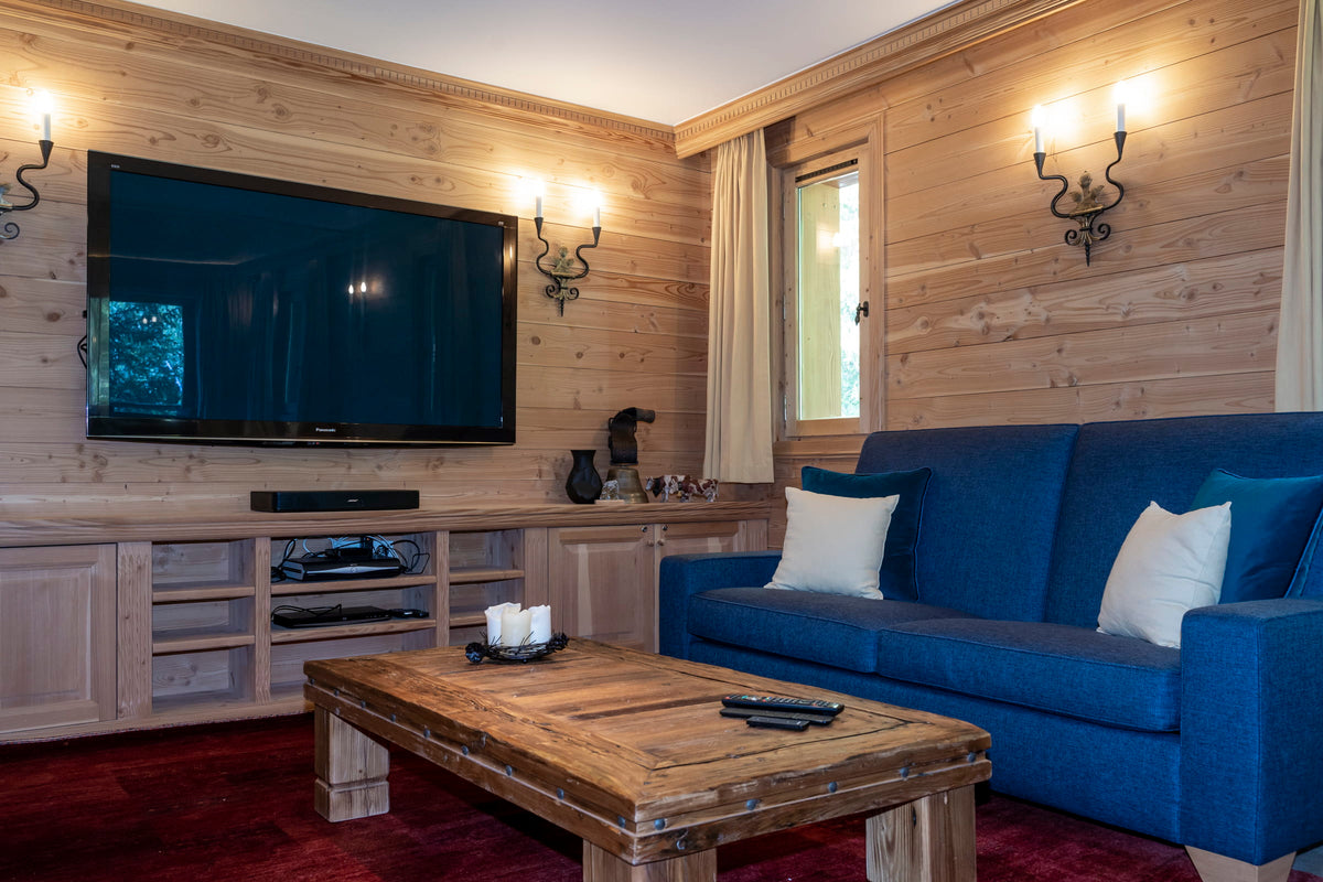 A cozy modern chalet-style living room featuring light wood paneled walls, a large black flat-screen TV mounted above a wooden media console, a blue fabric sofa with white pillows, a rustic wooden coffee table, and a deep red rug.