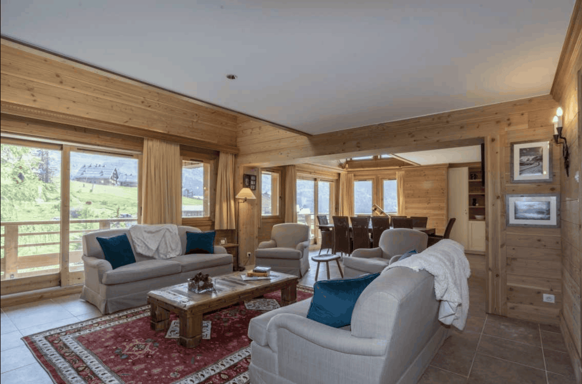A bright, modern chalet-style living room with light wood paneled walls and ceiling, featuring two light gray sofas with blue pillows, a wooden coffee table, and a red patterned rug. Large windows offer a view of green outdoor hills.