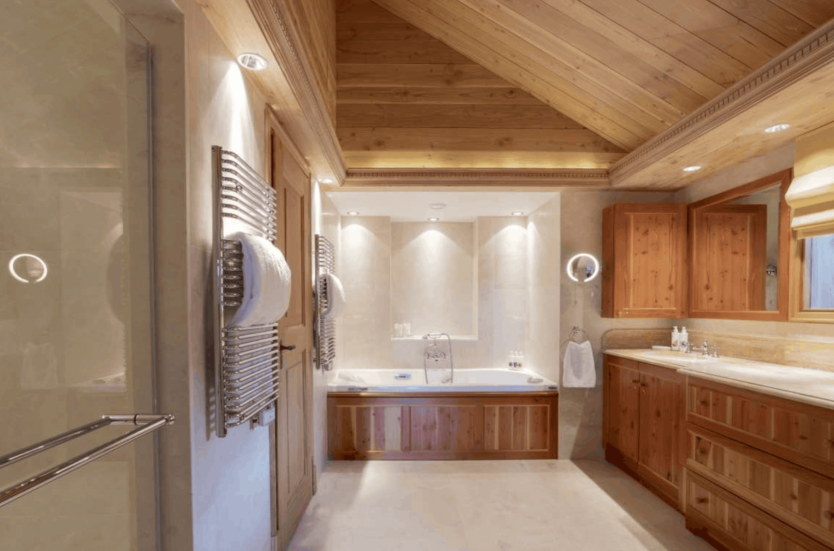 Luxury bathroom interior with warm wood cabinetry, a bathtub, a double vanity, and a glass shower door with a towel radiator on the left.
