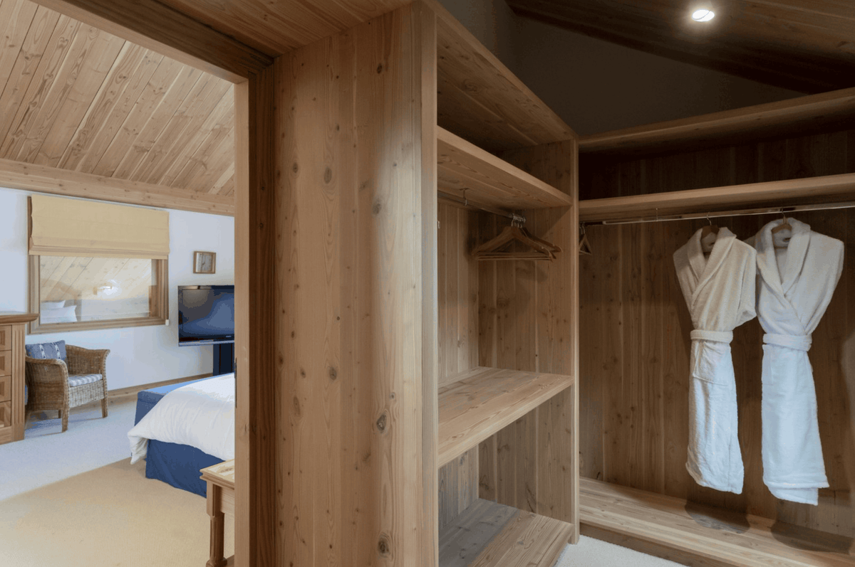 A rustic wooden walk-in closet with open shelving and two white bathrobes hanging on a hanger inside, shown in a bedroom setting.