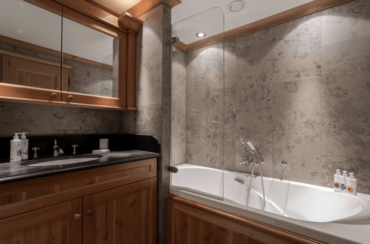 A luxury bathroom interior featuring a white bathtub with a glass shower screen, textured stone walls, and a wooden vanity with a black countertop and integrated mirror.