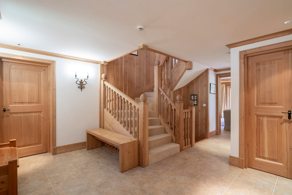 A home interior featuring a central wooden staircase with a wooden banister and a matching wooden bench at the base, flanked by light wood doors and beige tiled flooring.
