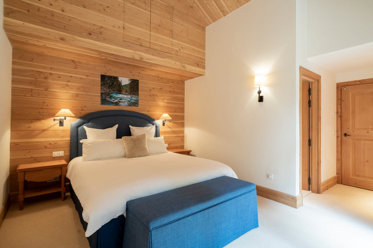 A modern bedroom featuring a double bed with white bedding and a dark blue upholstered headboard, set against a wood-paneled accent wall. A matching blue bench sits at the foot of the bed, with bedside tables, wall sconces, and a framed landscape print.