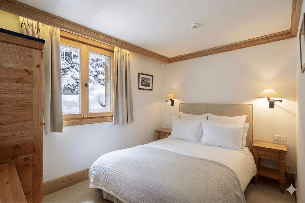 A cozy bedroom with a neatly made bed featuring white linens and a beige headboard, set against warm wood-paneled walls. A window on the left shows a snowy outdoor view, and two bedside tables with wall-mounted lamps flank the bed.