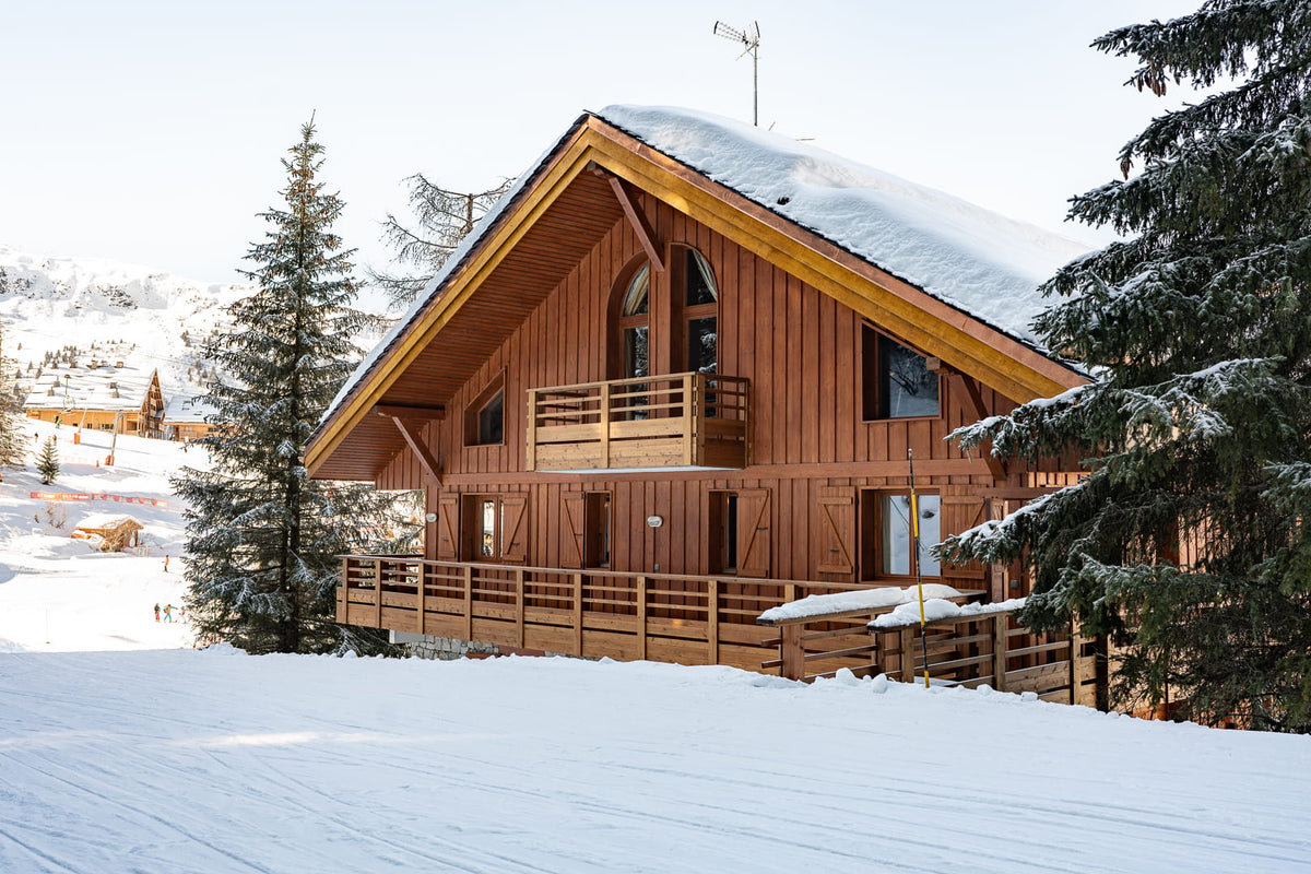 A wooden chalet-style mountain cabin with snow-covered roof and surrounding snowy landscape, framed by evergreen trees.