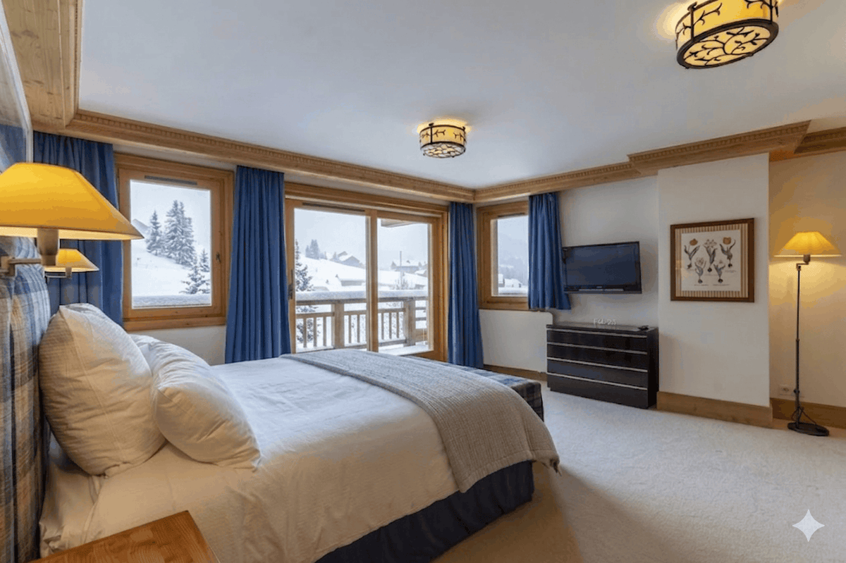 A luxury ski chalet bedroom featuring a large bed with white linens and blue plaid accents, large windows with blue curtains overlooking a snowy mountain landscape, a wall-mounted TV, and a floor lamp.