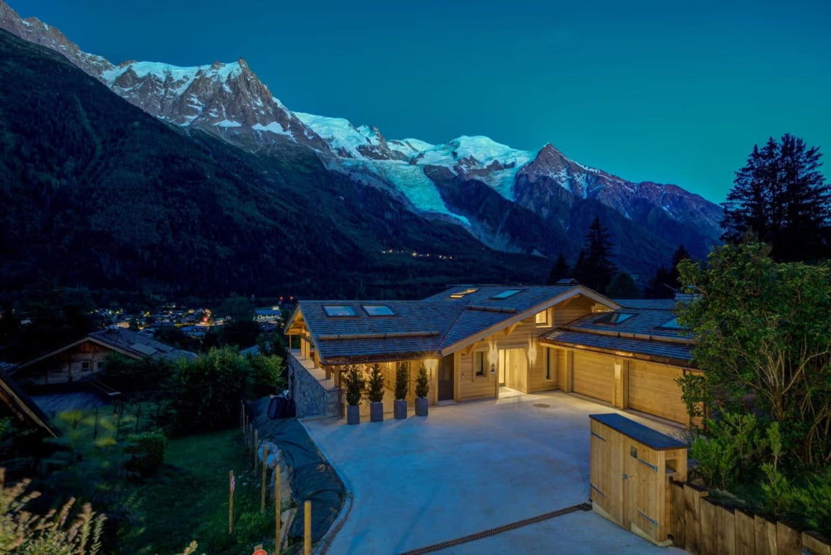 A luxury chalet-style mountain villa with warm glowing windows, set against a dramatic backdrop of snow-capped peaks under a deep blue twilight sky.