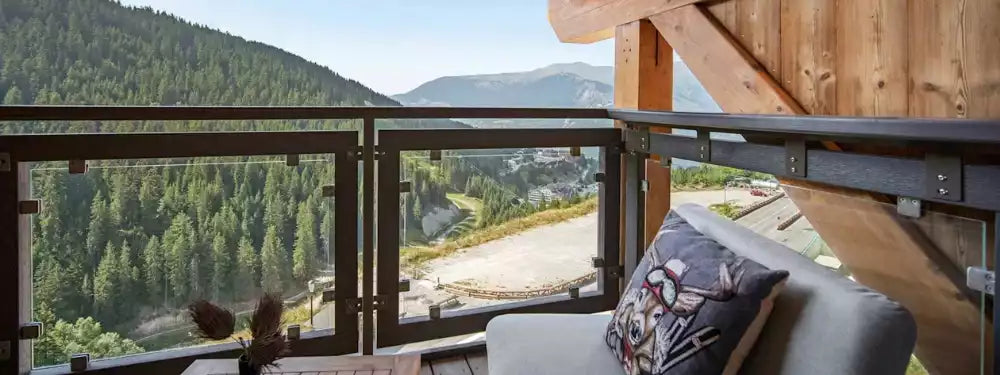 A modern outdoor balcony with glass panels and wooden framing, overlooking a lush green mountain forest valley. A gray lounge chair with a decorative pillow is placed on the balcony.