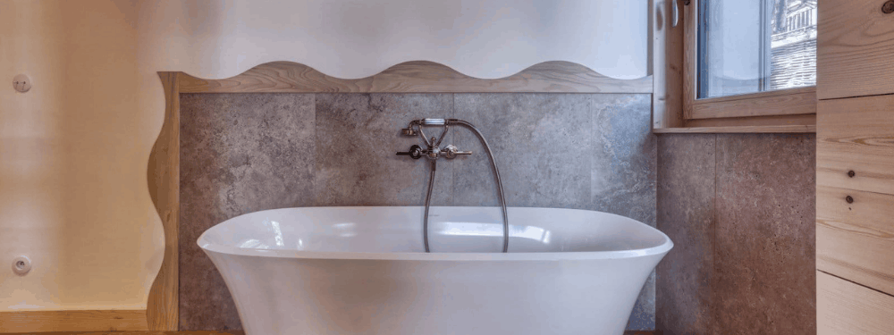 A freestanding white bathtub with a chrome faucet and handheld shower attachment, set against a gray stone tile wall with a decorative wavy wooden headboard and a window on the right.