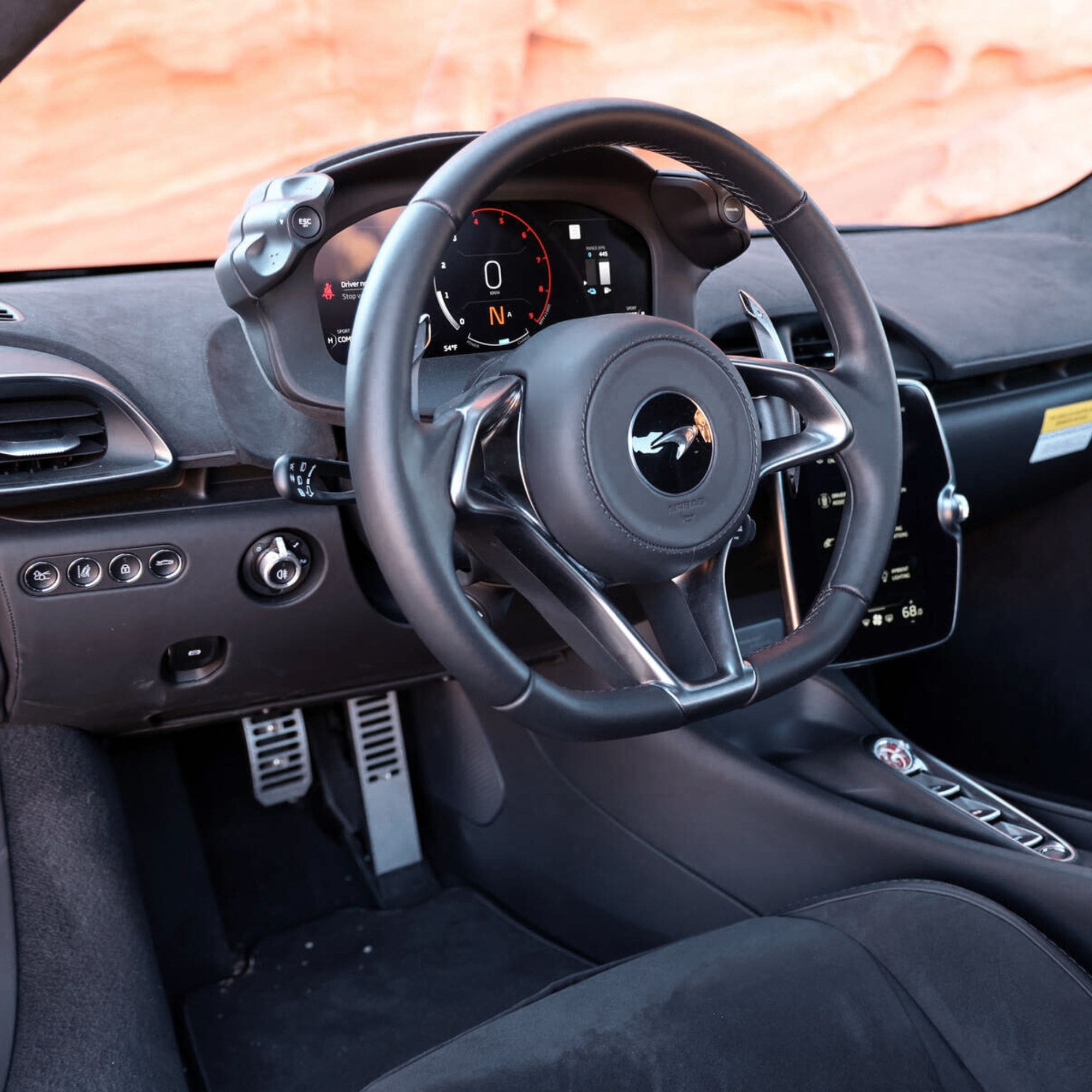 Close-up of a black leather steering wheel with a silver and black center logo, set inside a sports car cabin with a digital instrument cluster and a blurred orange rock landscape visible through the windshield.