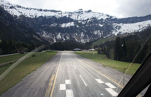 Runway in a mountainous area with snow-capped peaks and greenery.