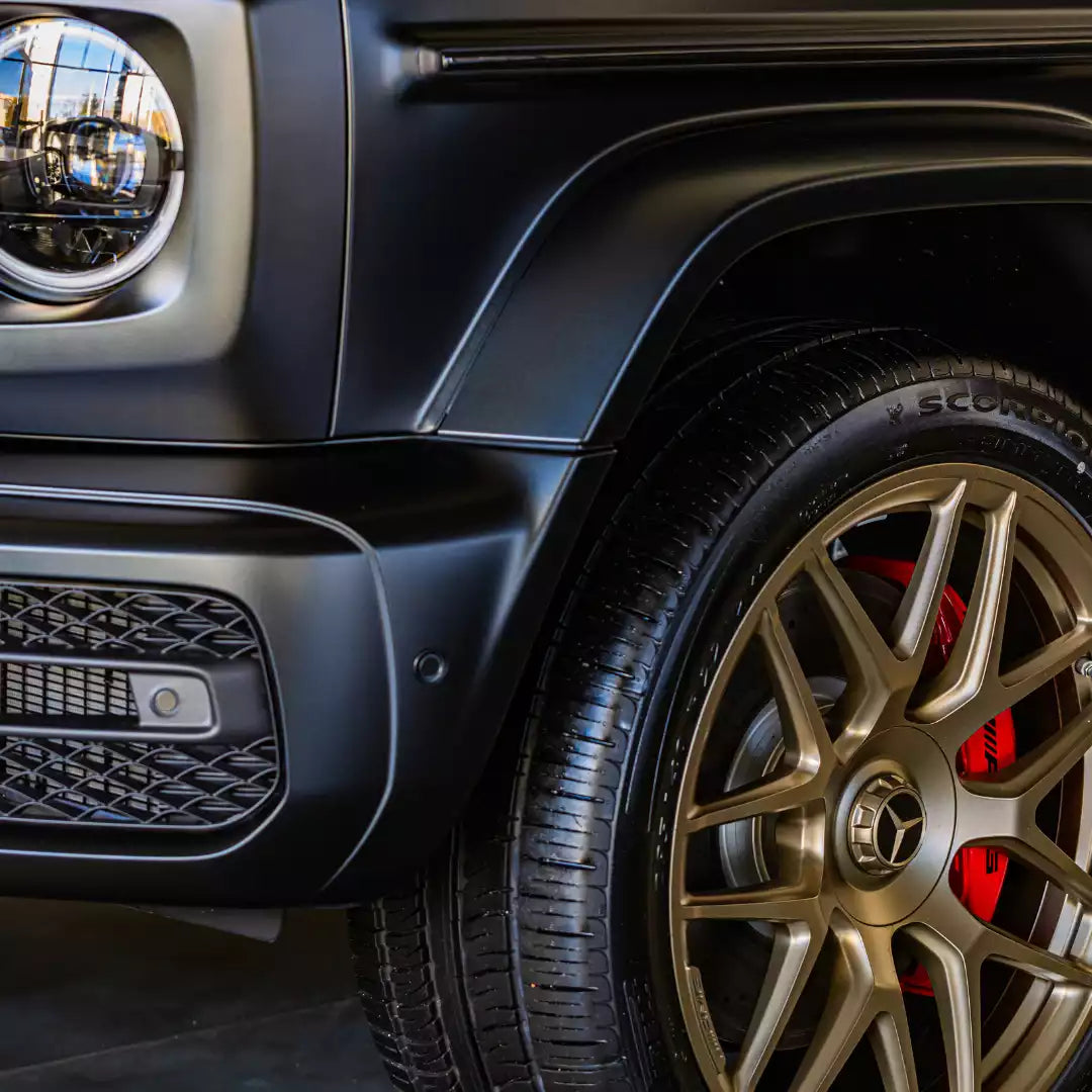 Close-up of a matte black Mercedes-Benz G-Class vehicle showing the front wheel, bronze multi-spoke alloy rim, red brake caliper, and textured tire sidewall.
