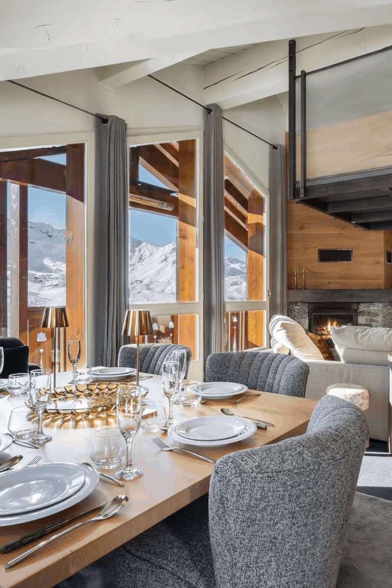A luxury modern chalet interior featuring a dining room with a wooden table set for six, gray upholstered chairs, large windows overlooking a snowy mountain landscape, and a loft bed in the background.